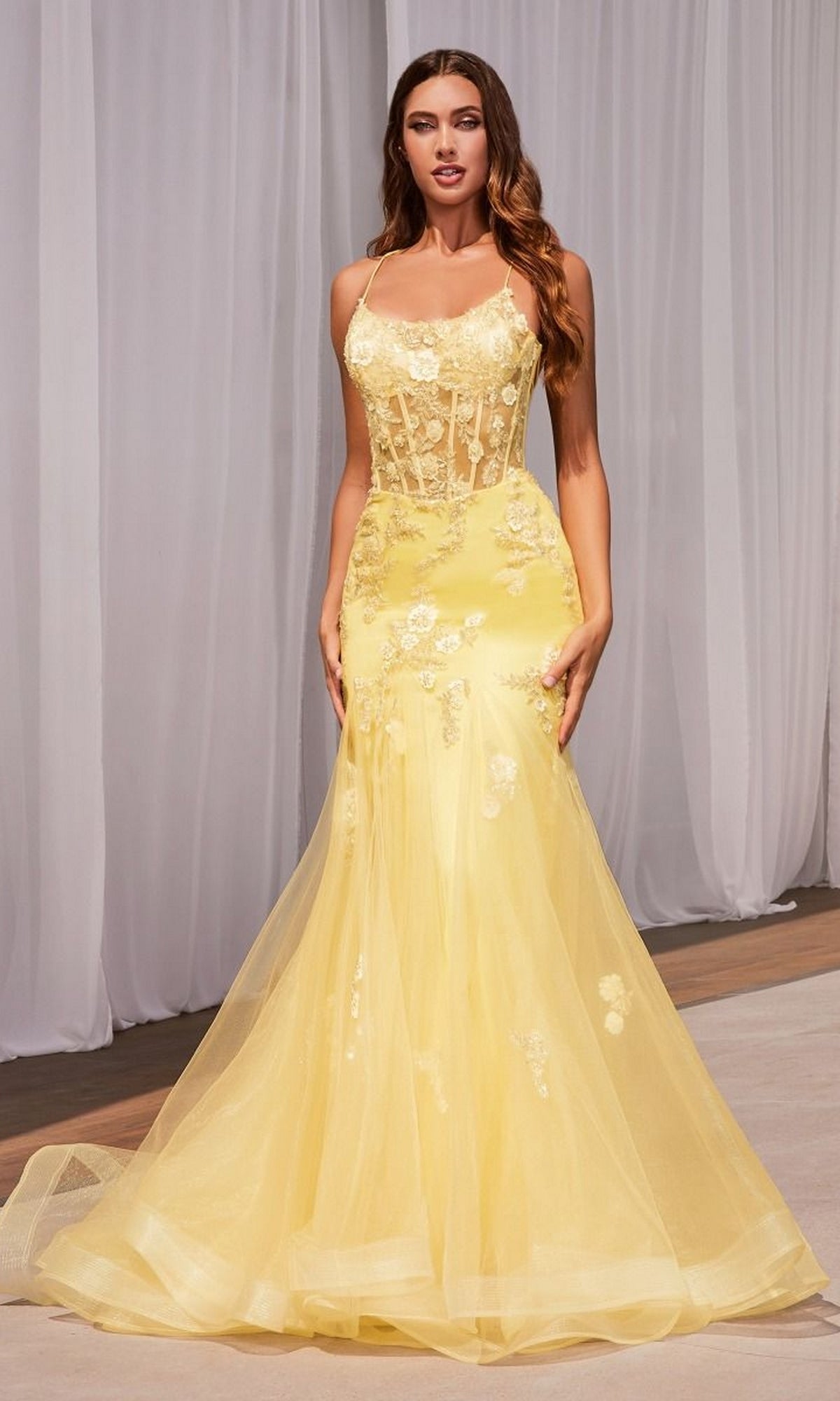 A woman stands indoors in the Long Pastel Floral Mermaid Prom Dress D145, a floor-length evening gown with a sheer, appliquéd bodice and delicate flowers. White curtains hang in the background behind her.